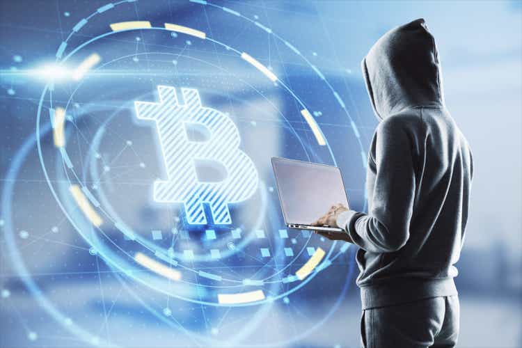 Hacker in hoodie using laptop computer with glowing bitcoin interface in blurry office interior. Hacking, theft and cryptocurrency concept. Double exposure.