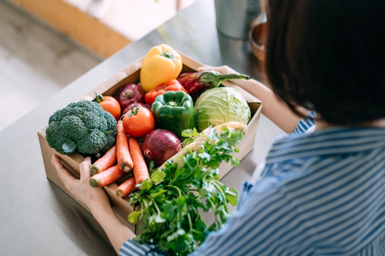 Over the shoulder view of young Asian woman holding a box full of colourful and fresh organic groceries in the kitchen counter at home