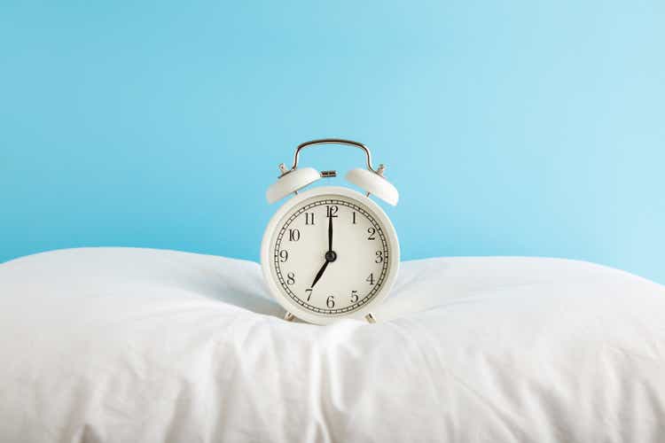White alarm clock on pillow at light blue wall background. Pastel color. 7 o"clock in morning. Waking time concept. Closeup. Front view.