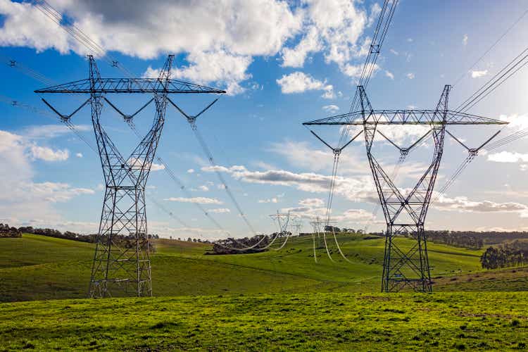 Double row of large electrical power pylons crossing lush green farmland with livestock grazing in the distance