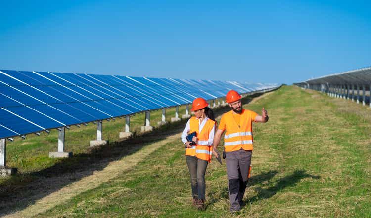 View of a solar power plant with two engineers walking and examining photovoltaic panels. Concept of alternative energy and its service
