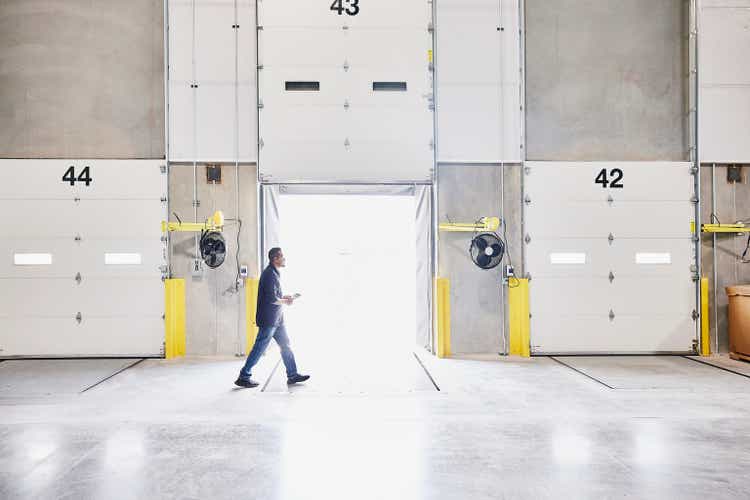 Wide shot of warehouse employee holding digital tablet and walking past open loading door in warehouse