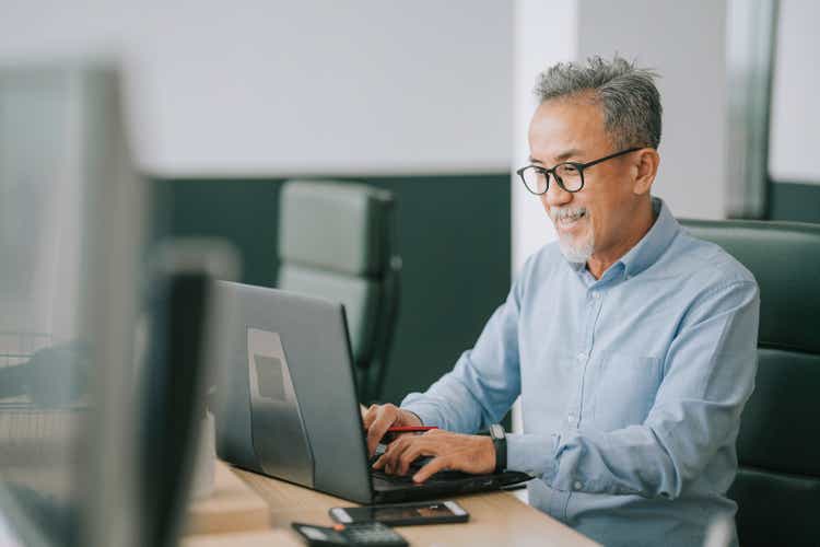 Asian chinese senior man with facial hair using laptop typing working in office open plan