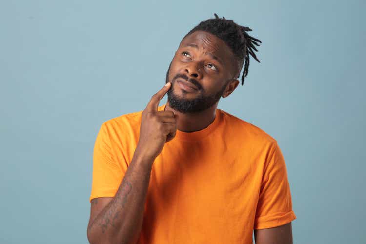 Portrait of male wearing orange t-shirt looking at his chin with hand. Thoughtful studio portrait against blue background