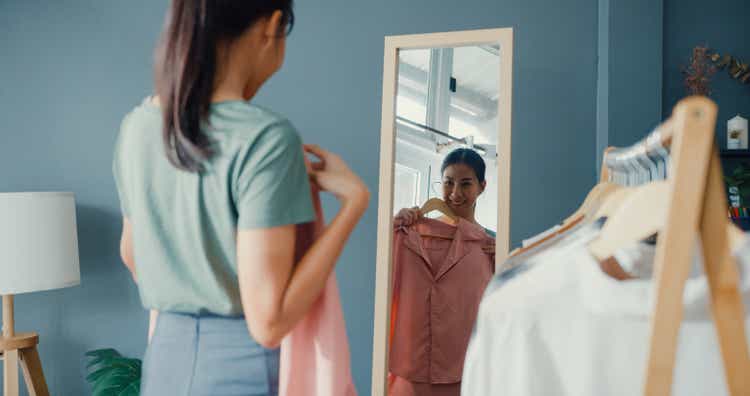 Beautiful attractive Asia lady choosing clothes on clothes rack dressing looking herself in mirror in living room at house.