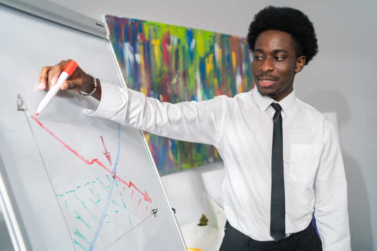 Handsome ambitious guy in white shirt tie with afro hair and stubble is standing in corporate office at white board, drawing diagram, charting company earnings promotions, conducting business training
