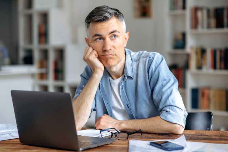 Middle aged caucasian business man or freelancer sitting at workplace, working on laptop, resting head on hand because of tired, doing work overtime, stressed and bored, need rest, pensive looks away