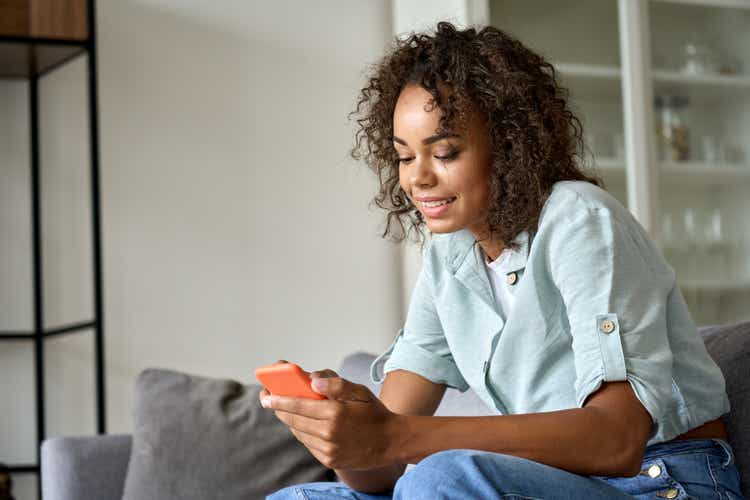 Smiling African American woman using mobile phone sitting on sofa at home.