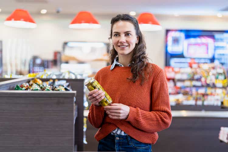 Portrait of smiling young woman holding olives jar while standing inside filling station store