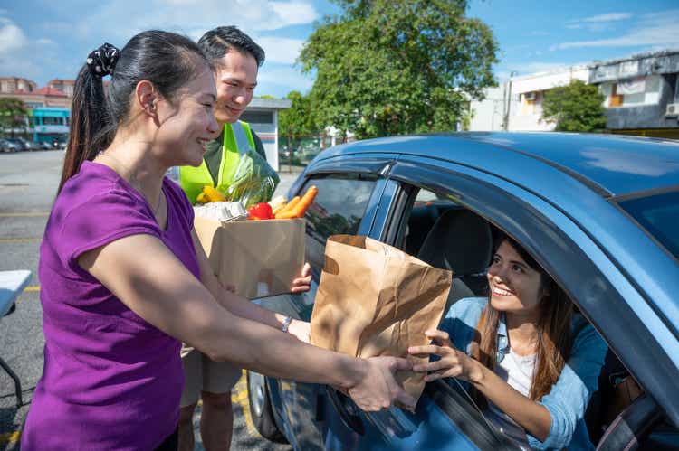 Young volunteers cheerfully hands a paper bag full of groceries for car driver through the car window.