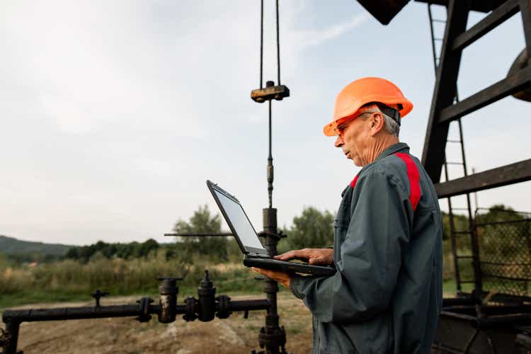 Oil worker checking work of the pumpjack in the oil well of the oil field using laptop