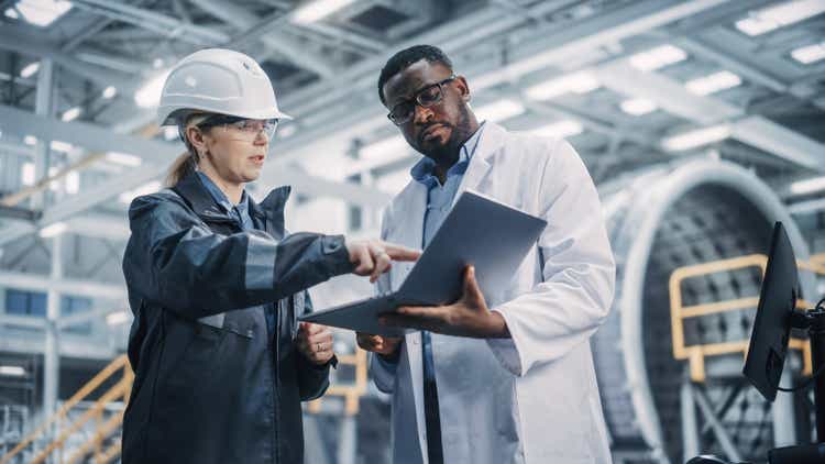 Team of Diverse Professional Heavy Industry Engineers Wearing Safety Uniform and Hard Hat Working on Laptop Computer. African American Technician and Female Worker Talking on a Meeting in a Factory.