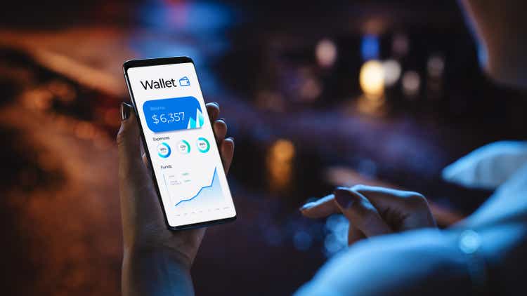 Beautiful woman using smartphone to check online wallet balance while standing on night city street full of neon lights.  Woman using the phone.  Close-up over the shoulder