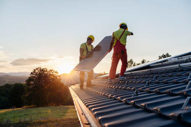 Workers Installing Solar Panels On Wooden House In Nature At Sunset.