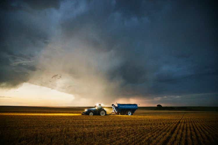 Wide shot of tractor pulling grain cart through cut wheat field ahead of storm clouds during summer harvest