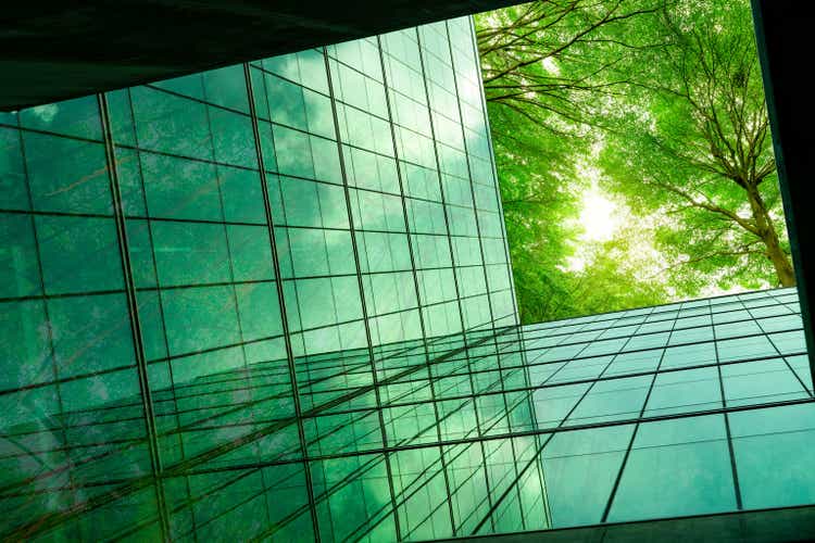 Eco-friendly building in the modern city. Green tree branches with leaves and sustainable glass building for reducing heat and carbon dioxide. Office building with green environment. Go green concept.