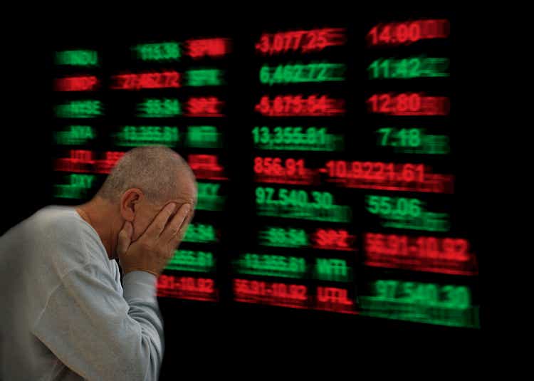 Unhappy man sitting with head in hands. In the background stock prices showing on a light board.