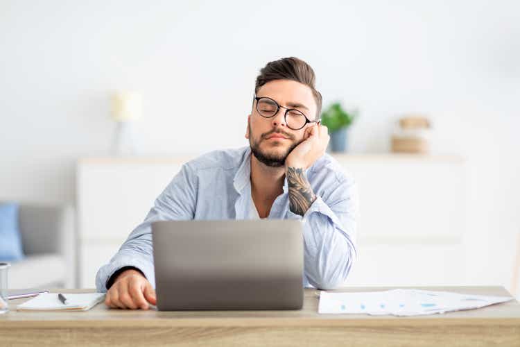 Overworked young freelancer man sitting at workplace and sleeping, napping while working on laptop, copy space
