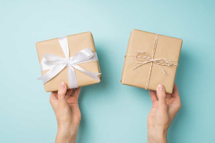 First person top view photo of female hands holding two craft paper gift boxes with twine bow and white satin ribbon bow on isolated pastel blue background