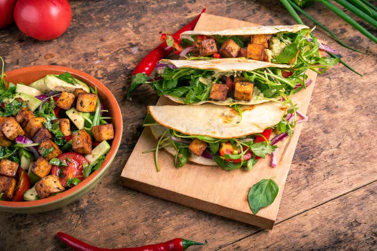 Tacos with vegetable salad and fried tofu on a wooden board on a table with a bowl of salad nearby