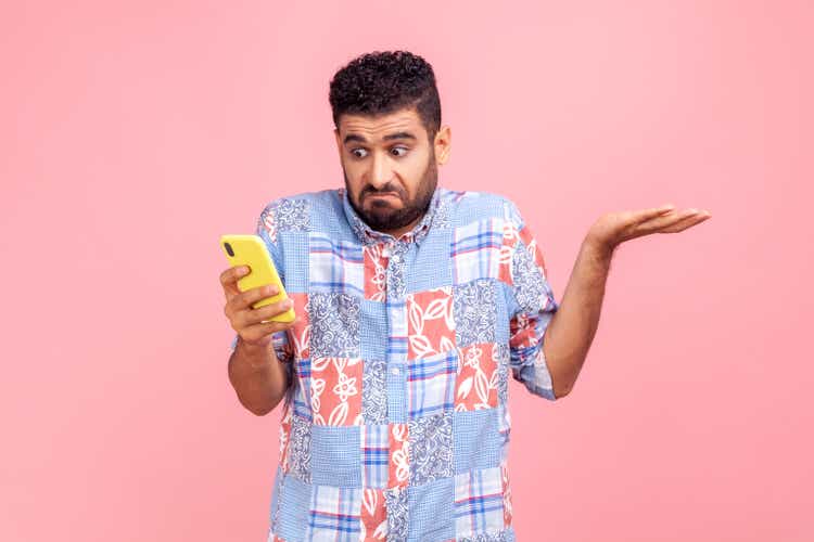 Portrait of attractive bearded man wearing blue casual shirt holding smart phone in hands, spreading hans aside, dont know how to use application.