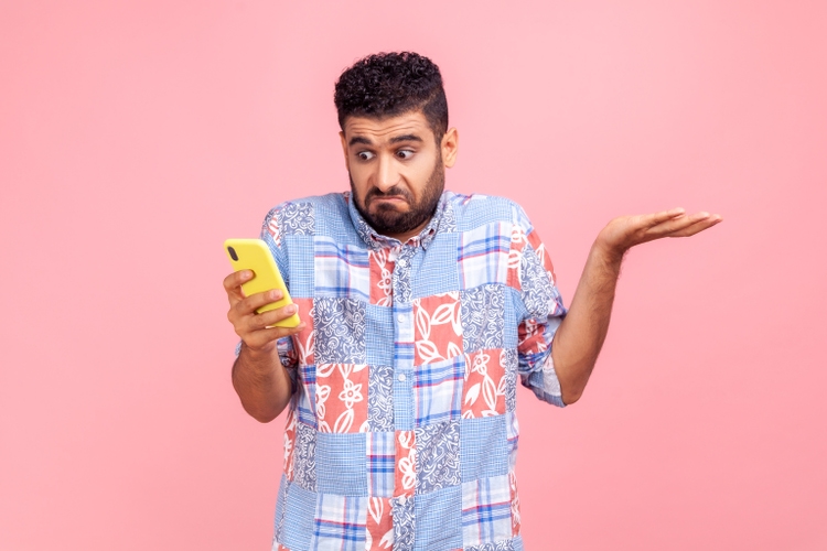 Portrait of attractive bearded man wearing blue casual shirt holding smart phone in hands, spreading hans aside, dont know how to use application.