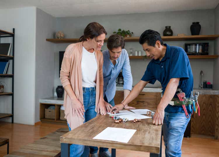 Plumber fixing a pipe and talking to his clients in the kitchen