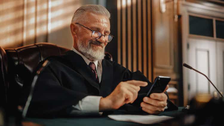 Court of Law and Justice on a Coffee Break: Smiling Male Judge Using Smartphone, Browsing Online Content, Using Social Media App, Investing, Internet e-commerce Purchases.