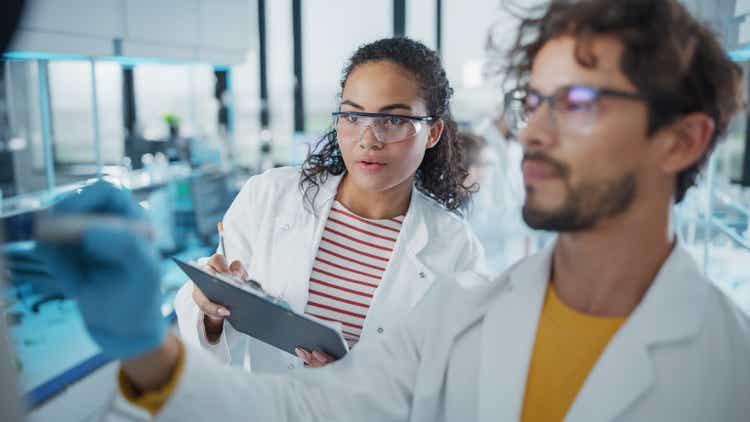 Medical Science Laboratory: Handsome Latin Male Scientist Writes Detailed Project Data Analysis on the Board, His Black Female Colleague Talks. Young Scientists Solving Problems.