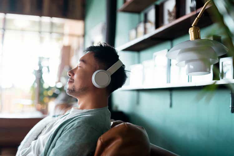 Young Asian man with eyes closed, enjoying music over headphones while relaxing on the sofa at home