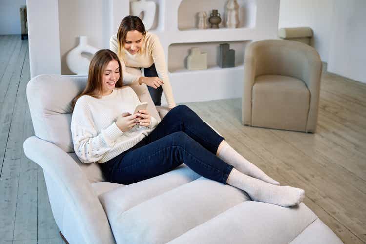 Two women on chaise longue with phone in lounge.