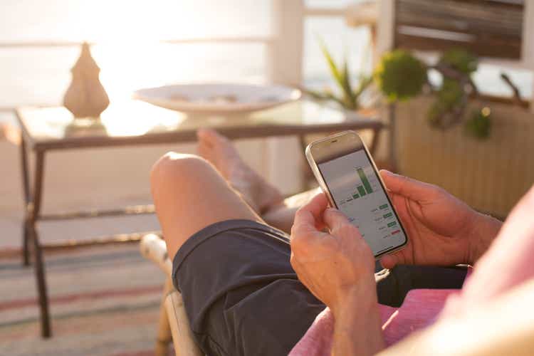 Man checking financial trading data on mobile phone while relaxing on terrace, close-up of hands