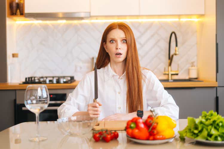 Portrait of attractive redhead young woman with shocked eyes holding big knife sitting at table with cutting board and looking at camera