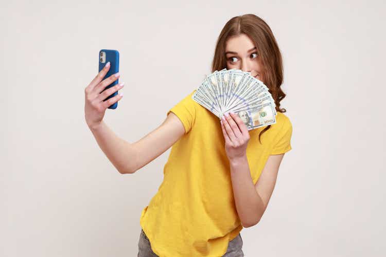 Portrait of rich teen girl with brown hair in T-shirt holding money and taking selfie by mobile phone, boasting of big profit, successful online betting.
