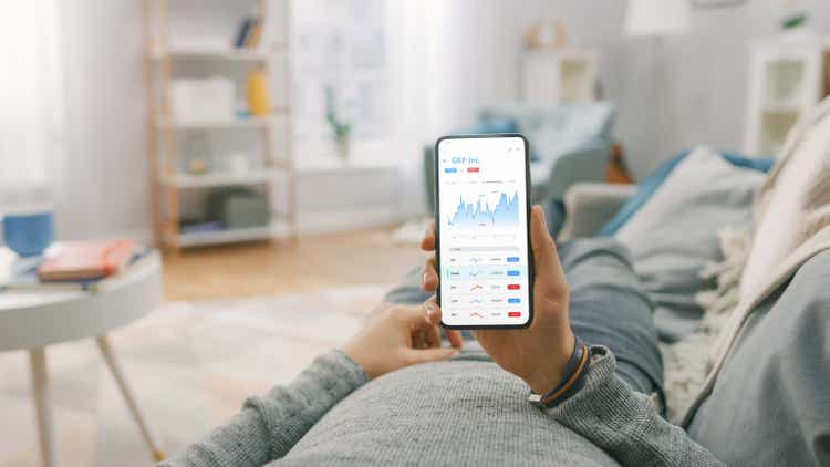 Man at Home Lying on a Couch and Using Smartphone with Stock Market App to Check Share Prices. Graph is Going Up Making a Great Profit. In the Background Cozy Homely Atmosphere.