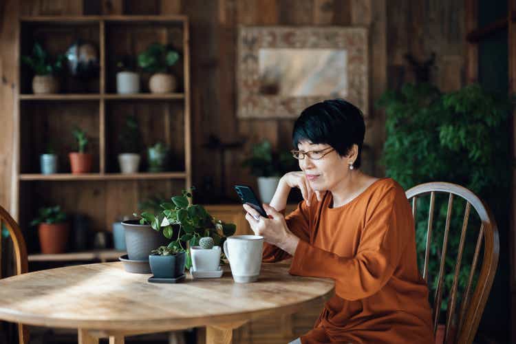 Smiling senior Asian woman sitting at the table, surfing on the net and shopping online on smartphone at home. Elderly and technology