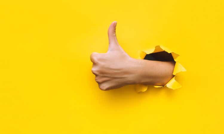The female hand shows a thumbs up gesture (like).  Torn hole in yellow paper.  Concept of positive attitude, approval and praise.  copy space.