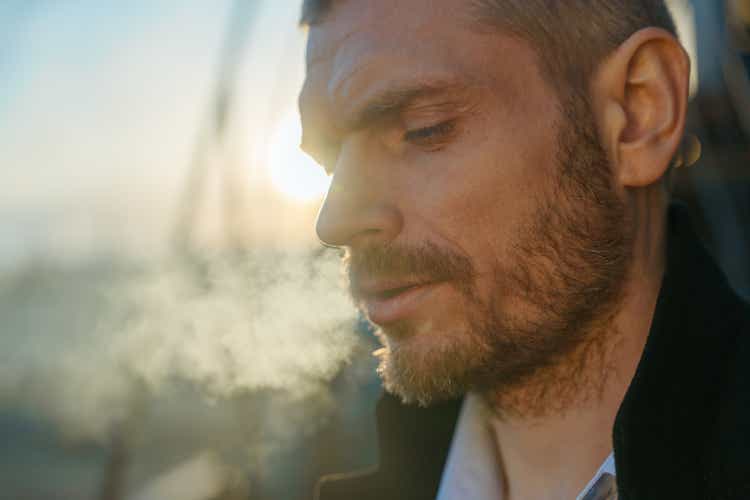 Close -up of a serious man smoking man looking down, standing on a roof during the dawn