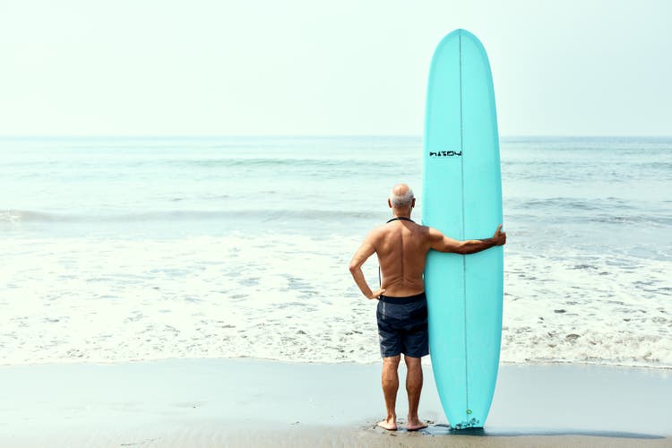 A portrait of a senior man in the back standing on the beach with a surfboard.