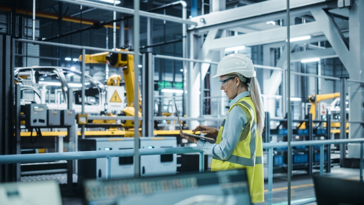 Car Factory: Female Automotive Engineer Wearing Hard Hat, Standing, Using Laptop. Monitoring, Control, Equipment Production. Automated Robot Arm Assembly Line Manufacturing Electric Vehicles.