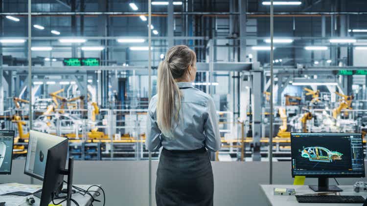 Bureau de l'usine automobile: Ingénieure en chef réussie supervisant le convoyeur de production de l'usine. Ligne d'assemblage de bras robotisés automatisés fabriquant des véhicules électriques de haute technologie avancés. Vue arrière Prise