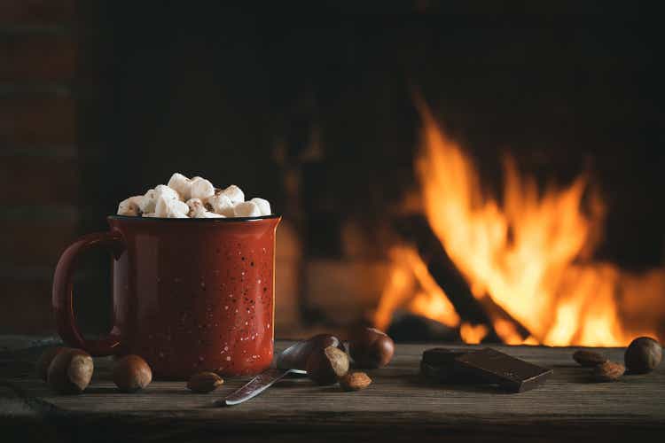 Cocoa with marshmallows and chocolate in a red mug on a wooden table near a burning fireplace