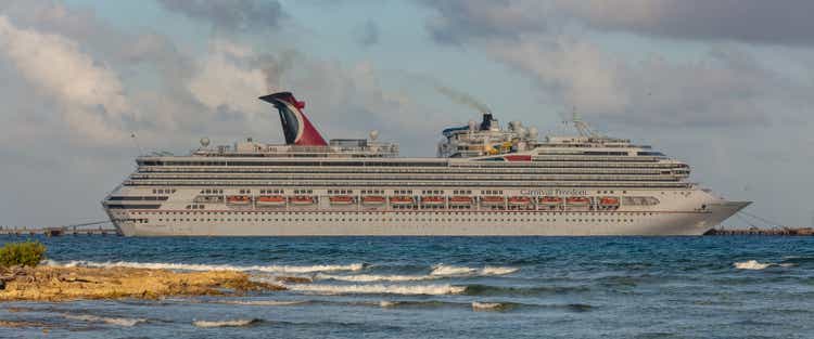 Beautiful panoramic shot of Carnival Freedom docked in Costa Maya port. Shore line, splashing waves in the foreground. Cloudy sky in the background