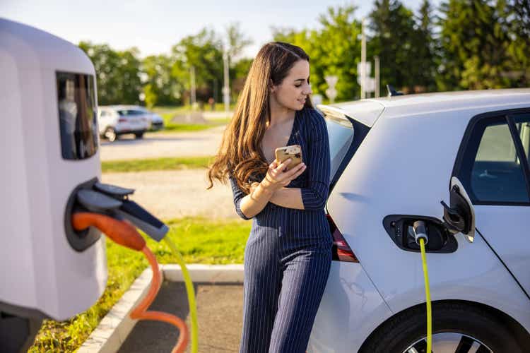 Young woman holding mobile phone while waiting for electric car to charge
