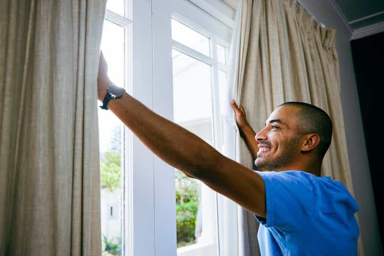 Shot of a young man opening up the curtains in a bedroom at home