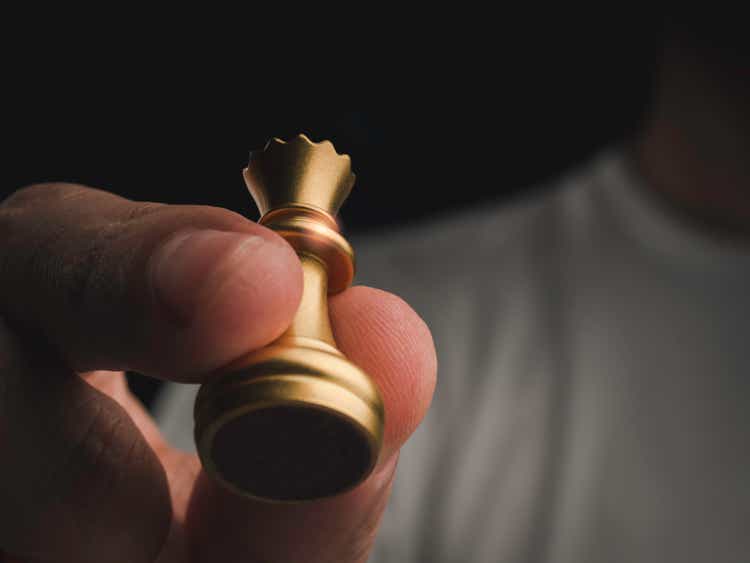 Close up gold queen chess piece in human hand in the air on dark background with copy space, below view. Hand moving queen chess figure on chess game competition. The winner Leadership concept.