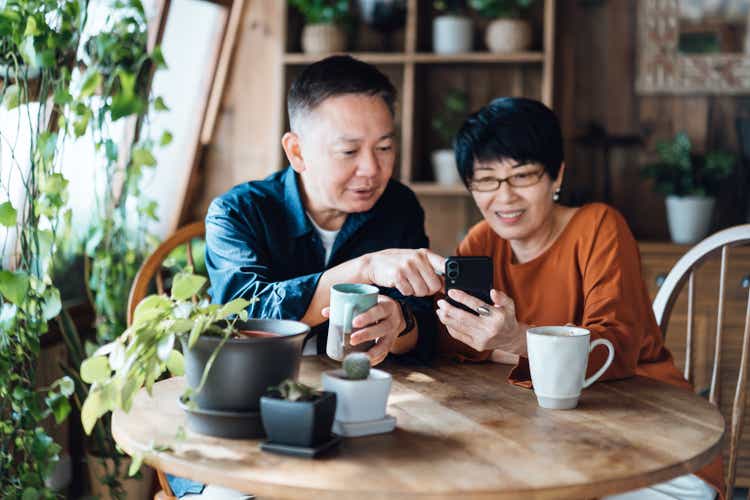 Happy senior Asian couple sitting at the table at home, managing online banking with mobile app on smartphone together. Accessing finances simply and securely. Senior lifestyle. Elderly and technology