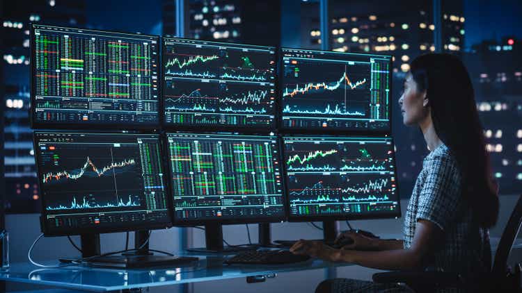 Financial Analyst Working on a Computer with Multi-Monitor Workstation with Real-Time Stocks, Commodities and Exchange Market Charts. Businesswoman at Work in Investment Broker Agency Office at Night.