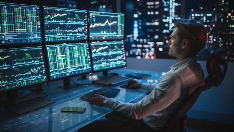 Financial Analyst Working on a Computer with Multi-Monitor Workstation with Real-Time Stocks, Commodities and Foreign Exchange Charts. Businessman Works in Investment Bank City Office Late Evening.