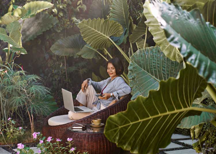 Asian woman working from home sitting in garden furniture surrounded by tropical plants, using laptop computer and mobile phone